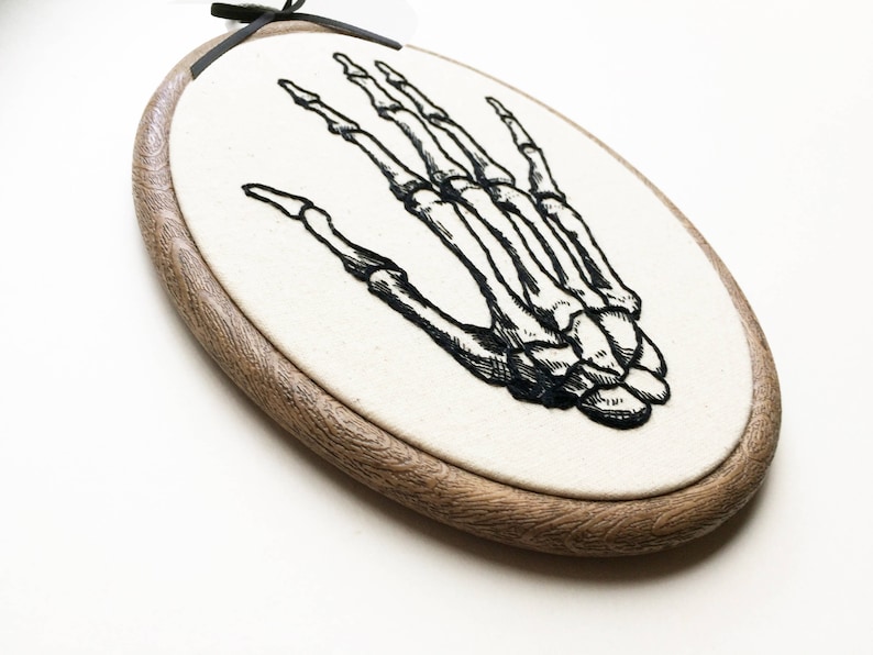 An oval embroidery hoop. A detailed embroidery of the bones of the hand is on the ivory fabric.