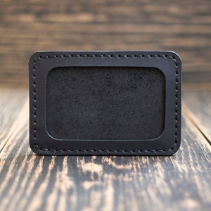 May include: A black leather card holder with a stitched border and a blank window for displaying identification.