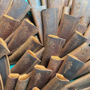 K&ouml;nnte beinhalten: Ein Stapel brauner Ledertaschen f&uuml;r Messer mit einem dekorativen Design aus drei Bl&auml;ttern. Die Taschen sind aus abgenutztem Leder mit einer dezenten Farbvariation in Braun und Beige.