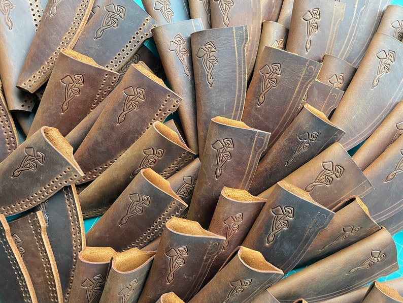 K&ouml;nnte beinhalten: Ein Stapel brauner Ledertaschen f&uuml;r Messer mit einem dekorativen Design aus drei Bl&auml;ttern. Die Taschen sind aus abgenutztem Leder mit einer dezenten Farbvariation in Braun und Beige.