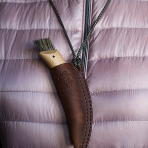 K&ouml;nnte beinhalten: Ein braunes Lederscheide mit einem Pilzdesign h&auml;ngt an einer Schnur um den Hals einer Person. Die Scheide enth&auml;lt eine B&uuml;rste mit Naturborsten und einem Holzgriff.