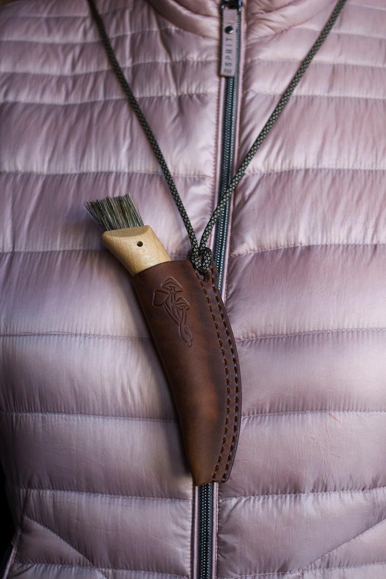K&ouml;nnte beinhalten: Ein braunes Lederscheide mit einem Pilzdesign h&auml;ngt an einer Schnur um den Hals einer Person. Die Scheide enth&auml;lt eine B&uuml;rste mit Naturborsten und einem Holzgriff.