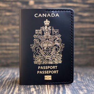 May include: A black passport holder with gold lettering and a detailed emblem. The word "CANADA" is at the top, with "PASSPORT PASSEPORT" below. The holder is made of a smooth material and has a stitched border.
