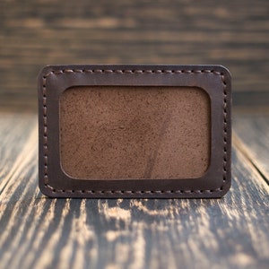 May include: A brown leather card holder with a stitched border and a clear plastic window for displaying identification.
