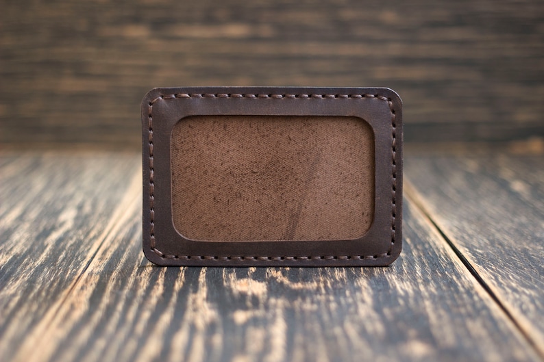 May include: A brown leather card holder with a stitched border and a clear plastic window for displaying identification.