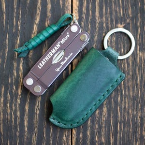 以下が含まれることがあります： 「Your Mechanic」の文字が入った茶色のLeatherman Microマルチツールと、緑色のレザーストラップ。緑色の革製シースとシルバーのキーリングも付属。濃い木材の表面に置かれたアイテム。