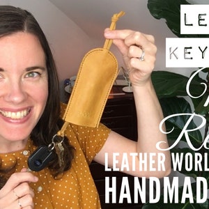 Op de afbeelding: Een vrouw houdt een gele leren sleutelhanger vast met een sleutelring en sleutels. De sleutelhanger is handgemaakt door Leather World. De tekst "LEATHER KEY CASE My Review LEATHER WORLD HANDMADE PLUS 10% OFF" staat op de achtergrond.