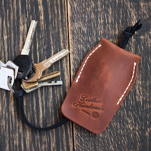 May include: Brown leather key holder with white stitching and a black cord. The holder has the text "Leather Handmade" embossed on it. A set of keys is attached to the cord.