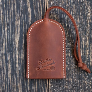 Op de afbeelding: Bruine leren sleutelhanger met witte stiksels en de tekst "Leather Handmades Est.2013" en een wereldbolpictogram.