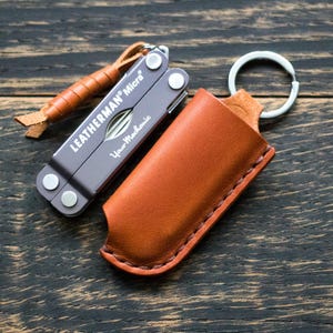 May include: A Leatherman Micra multi-tool with a brown leather sheath and a leather lanyard. The multi-tool is grey and silver, and the sheath is a warm brown colour. The text "Leatherman Micra" and "Your Mechanic" are visible.