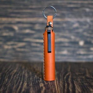 May include: A brown leather keychain with a silver key ring and a black metal clip. The keychain is designed to hold a small object, such as a lighter or a pen.