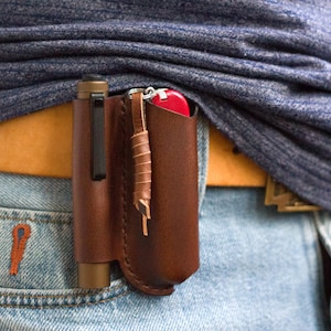 Puede incluir: Un organizador de cuero marrón montado en el cinturón que contiene un bolígrafo y una navaja de bolsillo roja. El organizador está sujeto a un cinturón de cuero marrón que se lleva con vaqueros azules. El bolígrafo es negro y dorado.