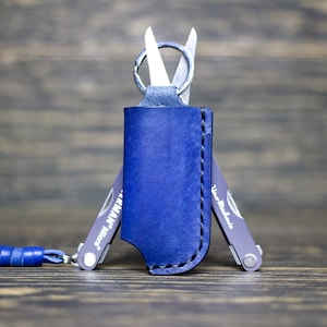 Könnte beinhalten: Ein blaues Lederetui mit einem silbernen Multitool, das an einem Schlüsselring befestigt ist. Das Multitool hat eine kleine Schere und der Text "Mini-Mizer" ist auf der Seite aufgedruckt.