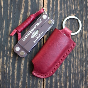 Könnte beinhalten: Ein Leatherman Micra Multitool mit einer roten Lederscheide und einem Schlüsselband. Das Multitool ist dunkelgrau mit dem Text "Leatherman Micra" und "Your Mechanic" bedruckt. Die Scheide ist rot und hat einen Schlüsselring.