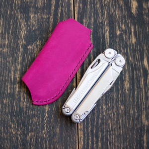 Op de afbeelding: Een zilverkleurige Leatherman Wave multitool in opgevouwen stand naast een fel magenta lederen etui. De multitool heeft een metalen afwerking met zichtbare schroeven en scharnieren, terwijl het etui gestikte randen en een voorgevormde bovenkant heeft.