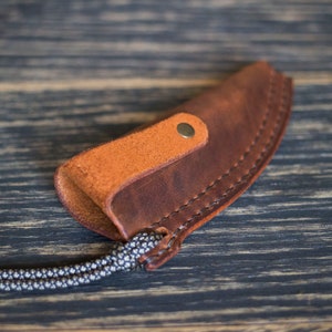 K&ouml;nnte beinhalten: Eine braune Ledertasche f&uuml;r ein Messer mit einer hellbraunen Ledertasche und einem Metall-Druckknopfverschluss. Die Tasche ist an einer geflochtenen Schnur befestigt.