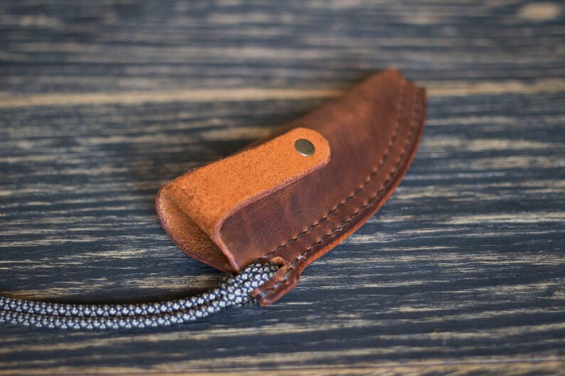 K&ouml;nnte beinhalten: Eine braune Ledertasche f&uuml;r ein Messer mit einer hellbraunen Ledertasche und einem Metall-Druckknopfverschluss. Die Tasche ist an einer geflochtenen Schnur befestigt.