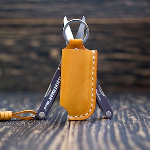 Könnte beinhalten: Ein braunes Lederetui mit weißem Steppdesign enthält ein kleines Multitool mit einem Schlüsselring. Das Multitool ist ein Leatherman Micra.