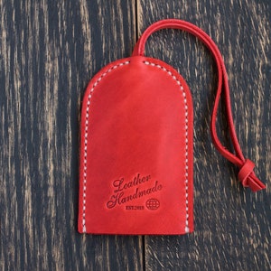 Op de afbeelding: Rode leren sleutelhanger met een witte gestikte rand. De sleutelhanger heeft een lus om sleutels aan te bevestigen en heeft de tekst "Leather Handmade Est. 2015" met een wereldbolpictogram.