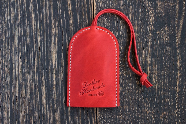 Op de afbeelding: Rode leren sleutelhanger met een witte gestikte rand. De sleutelhanger heeft een lus om sleutels aan te bevestigen en heeft de tekst "Leather Handmade Est. 2015" met een wereldbolpictogram.