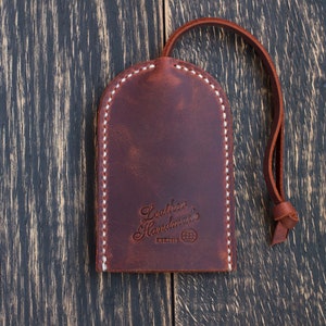 Op de afbeelding: Een bruine leren sleutelhanger met een witte gestikte rand. De sleutelhanger heeft een lus om sleutels aan te bevestigen en de tekst "Leather Handmade" met een wereldbol symbool en het woord "ESTABLISHED" eronder.