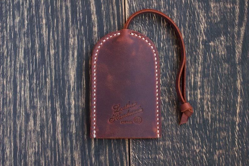 Op de afbeelding: Een bruine leren sleutelhanger met een witte gestikte rand. De sleutelhanger heeft een lus om sleutels aan te bevestigen en de tekst "Leather Handmade" met een wereldbol symbool en het woord "ESTABLISHED" eronder.
