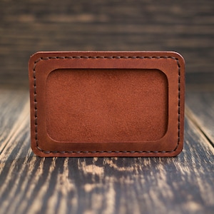 May include: A brown leather card holder with a stitched border and a blank rectangular space in the center.