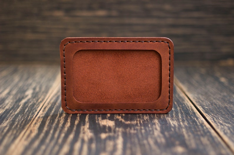 May include: A brown leather card holder with a stitched border and a blank rectangular space in the center.