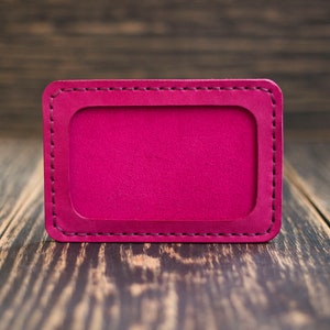 May include: A bright pink leather card holder with a stitched border. The card holder is on a wooden surface.