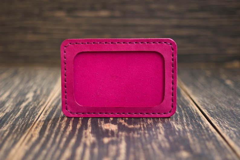 May include: A bright pink leather card holder with a stitched border. The card holder is on a wooden surface.