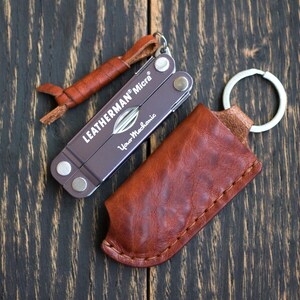 Könnte beinhalten: Ein Leatherman Micra Multitool mit einer braunen Lederscheide und einem kleinen Lederanhänger. Das Multitool ist dunkelgrau mit dem Markennamen und "Your Mechanic" aufgedruckt. Die Scheide ist braun und hat einen Schlüsselring.