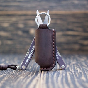 Könnte beinhalten: Ein braunes Lederetui mit einem genähten Rand enthält ein kleines Multitool mit einem Schlüsselring. Das Multitool ist ein Leatherman Micra und hat eine silberne Oberfläche. Das Etui und das Multitool befinden sich auf einer Holzoberfläche.