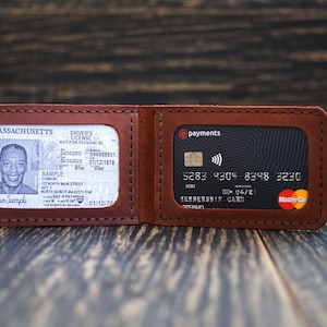 Puede incluir: Una cartera de cuero marrón con dos ranuras para tarjetas. La ranura izquierda contiene una licencia de conducir de Massachusetts con el nombre "Coma Sample" y la ranura derecha contiene una tarjeta de crédito negra con el texto "@payments" y "MEMBERSHIP CARD".