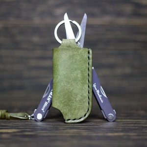 May include: A green leather case containing a small multi-tool. The multi-tool features a small blade, a nail clipper, and a small screwdriver. The case has a loop for attaching to a keychain.