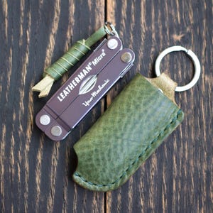 Könnte beinhalten: Ein Leatherman Micra Multi-Tool mit einer grünen Lederscheide und einem kleinen, grün umwickelten Werkzeug. Das Multi-Tool ist lila mit dem Text "Leatherman Micra" und "Your Mechanic". Die Artikel liegen auf einer dunklen Holzoberfläche.