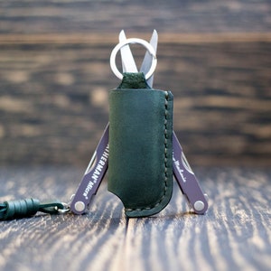 Könnte beinhalten: Ein grünes Lederetui mit einem kleinen Multitool darin. Das Multitool hat einen Schlüsselring und der Text "Leatherman Micra" ist an der Seite sichtbar.