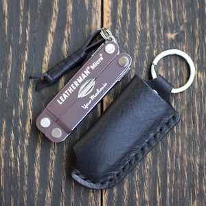 Könnte beinhalten: Ein Leatherman Micra Multi-Tool mit einer schwarzen Lederscheide und einem Schlüsselring. Das Multi-Tool ist lila mit dem Text "LEATHERMAN Micra" und "Your Mechanic". Die Scheide ist schwarz und hat einen genähten Rand.