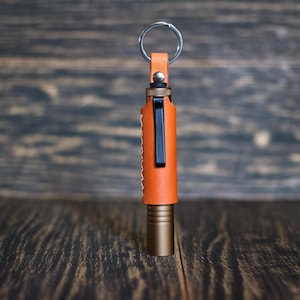 Może przedstawiać: Pomarańczowa latarka owinięta skórą z czarnym klipsem i metalowym brelokiem. Latarka ma podstawę w kolorze brązu i małą skórzaną pętlę na górze. Tło stanowi ciemna drewniana powierzchnia.