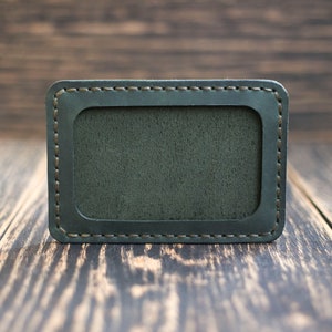 May include: A dark green leather card holder with a stitched border and a rectangular cutout for a card. The holder is on a wooden surface.
