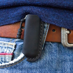 May include: A black leather sheath with visible stitching and a black cord pull tab, clipped onto a brown leather belt worn over blue denim jeans. The sheath is vertically oriented and features a smooth, matte finish on the exterior surface.