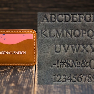 May include: A brown leather card holder with a pink insert and the word "PERSONALIZATION" embossed on the front. A sample alphabet and numbers are shown on a separate piece of leather.