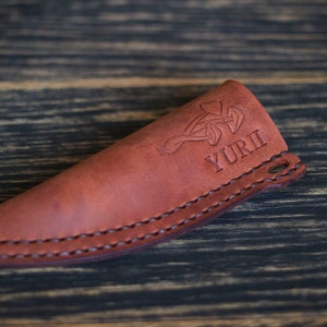 K&ouml;nnte beinhalten: Eine braune Ledertasche f&uuml;r ein Messer mit einem keltischen Knotendesign und dem eingepr&auml;gten Namen "YURI".