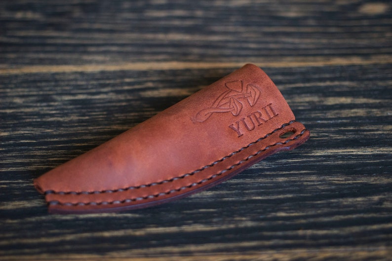 K&ouml;nnte beinhalten: Eine braune Ledertasche f&uuml;r ein Messer mit einem keltischen Knotendesign und dem eingepr&auml;gten Namen "YURI".