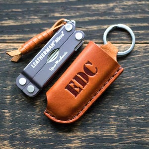 Könnte beinhalten: Ein Leatherman Micro Multi-Tool und eine braune Leder-EDC-Tasche (Everyday Carry) mit Schlüsselring. Das Multi-Tool ist grau mit dem Text "Leatherman Micro" und "Your Mechanic". Die Ledertasche hat die Buchstaben "EDC" ausgeschnitten.