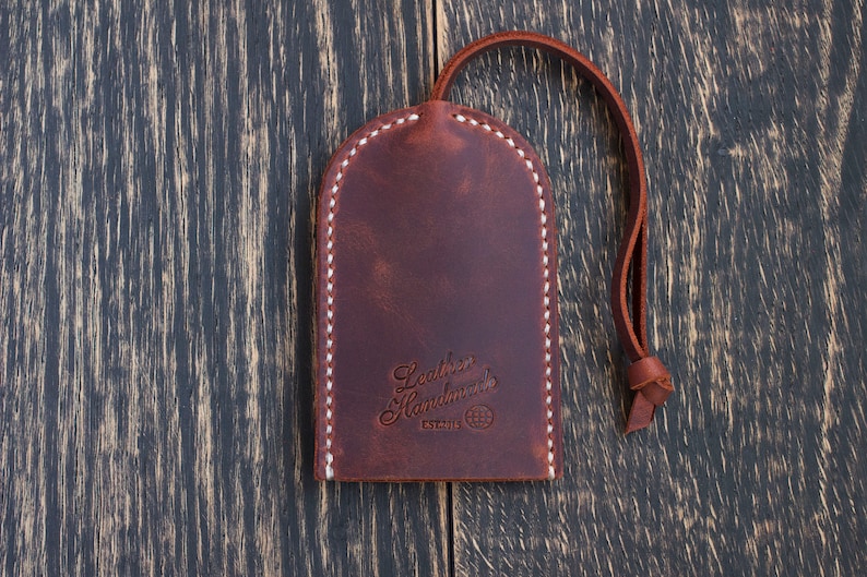 May include: A brown leather key chain with a white stitched border. The keychain has a loop for attaching keys and the text "Leather Handmade" with a globe symbol and the word "ESTABLISHED" below it.