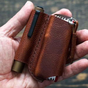 Pode incluir: Um porta-ferramentas múltiplo de couro castanho com um clipe preto e um acessório de lanterna de cor latão. O suporte é costurado e tem um fecho de correr. O couro tem um aspeto texturizado.