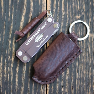 Può includere: Un multiutensile Leatherman Micra con un fodero in pelle marrone e un cordino in pelle. Il multiutensile è di colore viola tenue con il testo "Leatherman Micra" e "Your Mechanic" stampato sopra. Il fodero è marrone scuro.