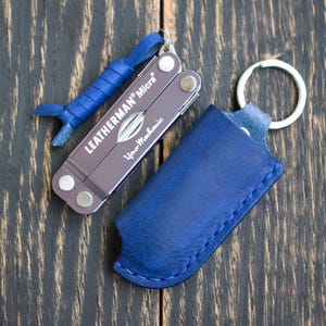 Könnte beinhalten: Ein Leatherman Micra Multi-Tool mit einer blauen Lederscheide und einem blauen Leder-Schlüsselanhänger. Das Multi-Tool ist braun mit silbernen Akzenten und dem Text "Leatherman Micra" und "Your Mechanic". Auf dunkler Holzoberfläche.