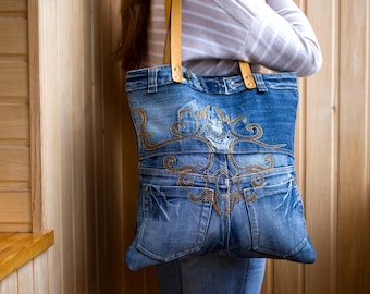 May include: A blue denim tote bag with a brown leather strap and gold embroidery. The bag is made from recycled denim and has a unique design.