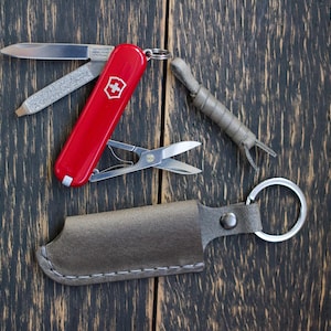 Puede incluir: Una navaja suiza roja con múltiples herramientas, incluyendo una hoja, tijeras y lima, junto a una funda de cuero y una pequeña herramienta gris. Los artículos están dispuestos sobre una superficie de madera oscura.